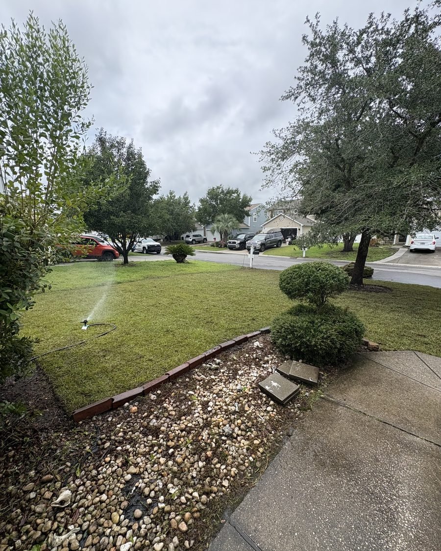 A sprinkler head and river rock bed along a sidewalk beside a freshly watered lawn
